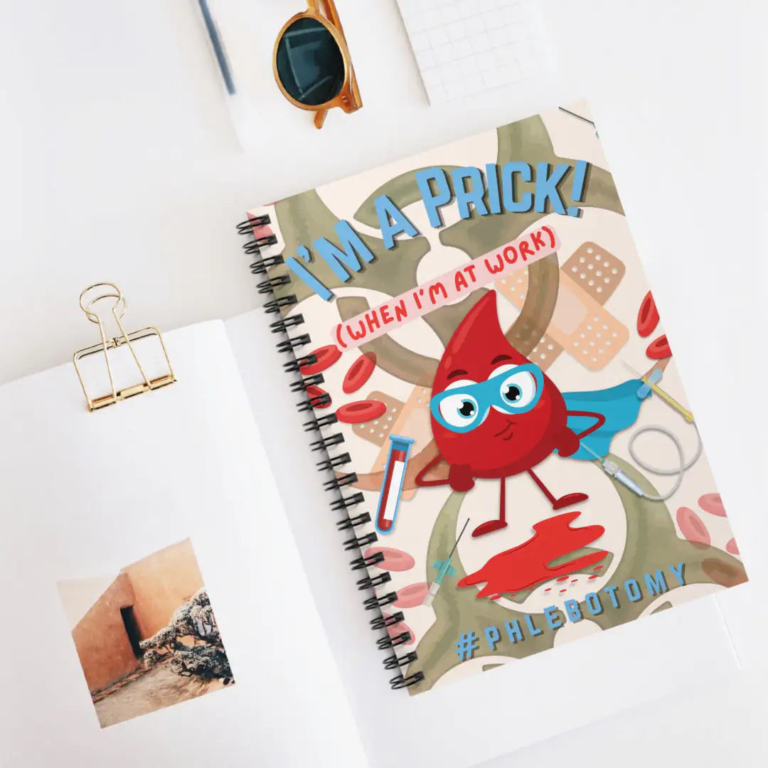 Notebook with a cartoon blood cell design and text 'I'm A Prick When I'm At Work' on a white desk.