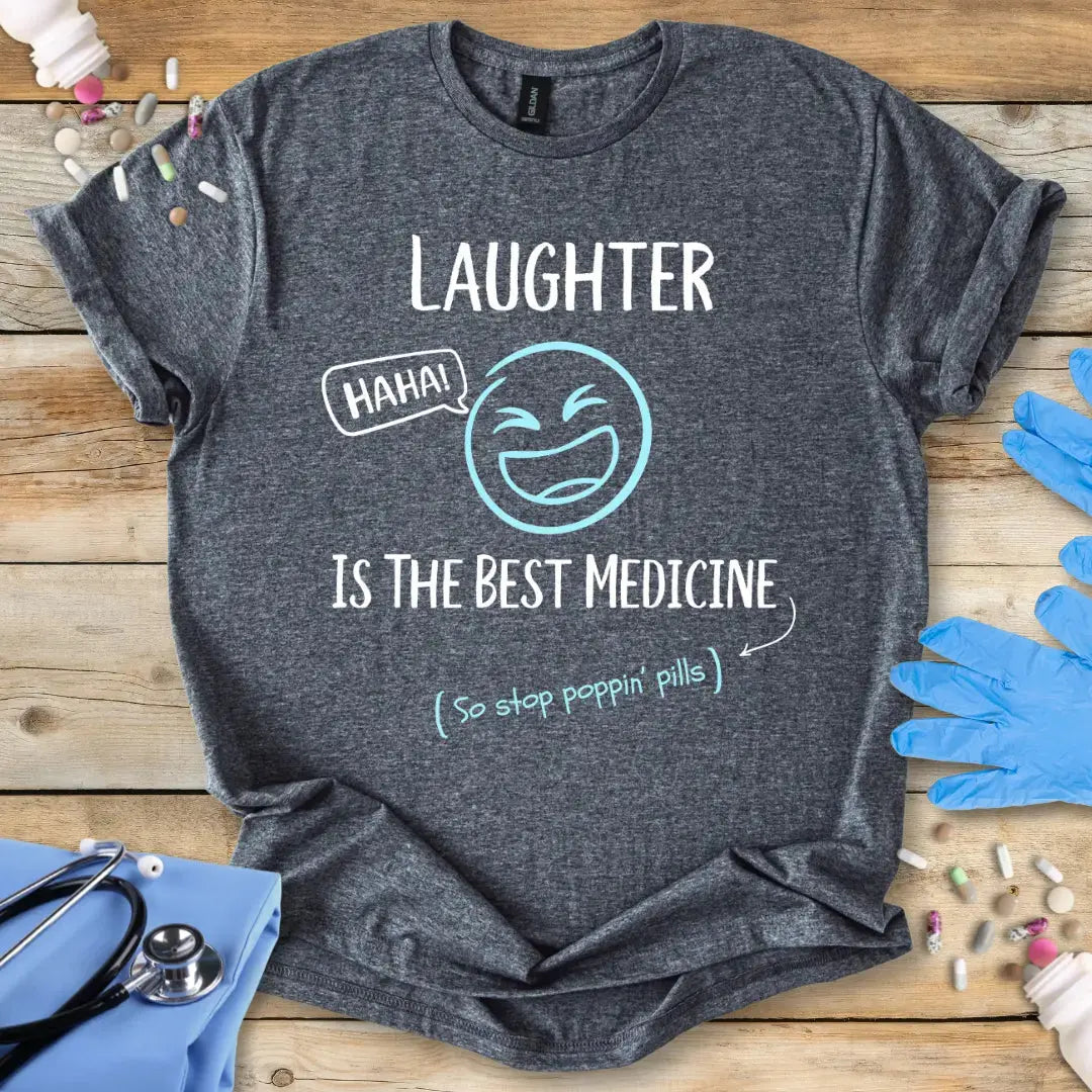 Gray-t-shirt with medical humor text 'Laughter Is The Best Medicine' and graphics on a wooden surface with medical-themed items.