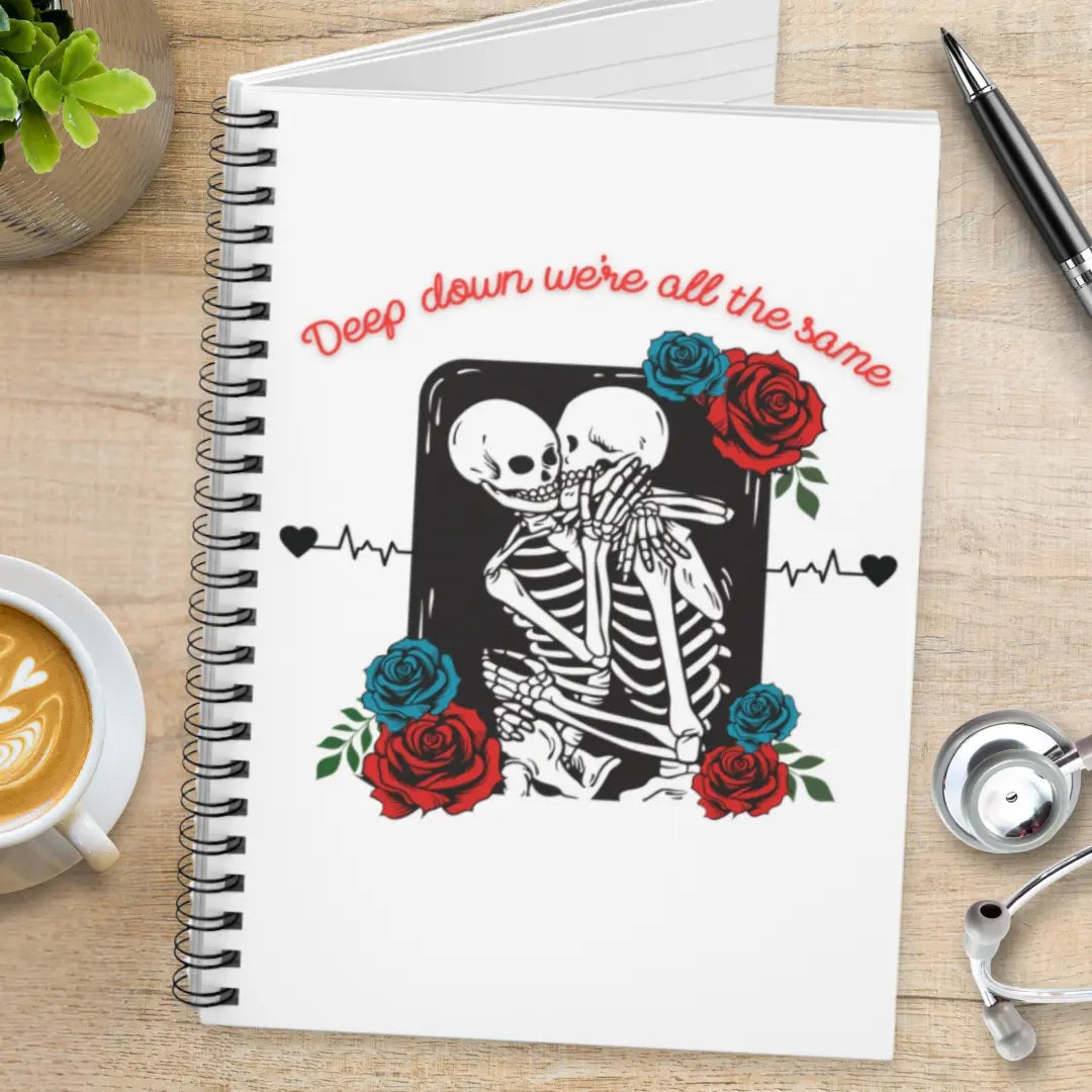 Notebook with skeleton design and roses and' Deep Down We're All The Same' text on a wooden surface with coffee and a pen.