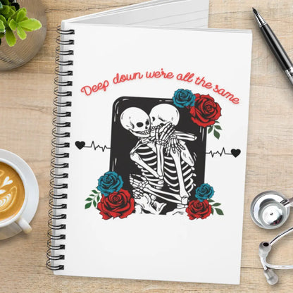 Notebook with skeleton design and roses and' Deep Down We're All The Same' text on a wooden surface with coffee and a pen.