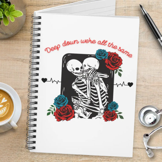Notebook with skeleton design and roses and' Deep Down We're All The Same' text on a wooden surface with coffee and a pen.