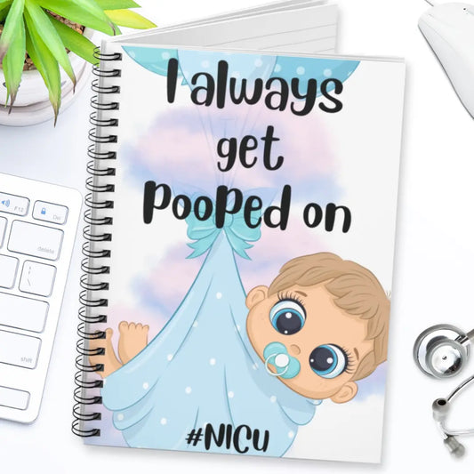 Notebook with cartoon baby illustration and 'I Always Get Pooped On' text on a desk with stethoscope.