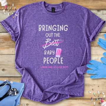 Purple t-shirt with humorous 'Bringing Out The Baby In People' text on a wooden surface with medical-themed items.