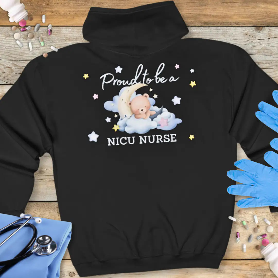 Black hoodie back printed with 'Proud to be a NICU Nurse' in script and bold text, featuring a watercolor teddy bear resting on clouds with a crescent moon and colorful stars, on a wooden background with medical props.