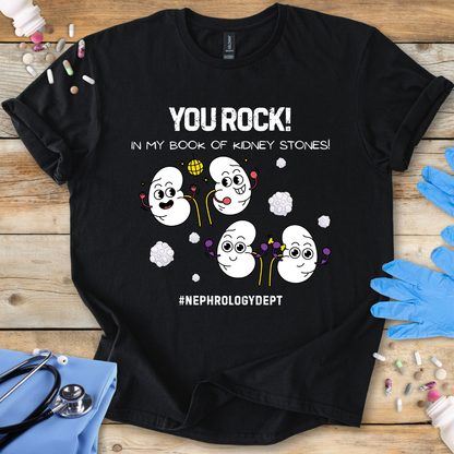Black t-shirt with cartoon kidneys and 'You rock in my book of kidney stones, nephrology dept.' text on a wooden surface with medical accessories.
