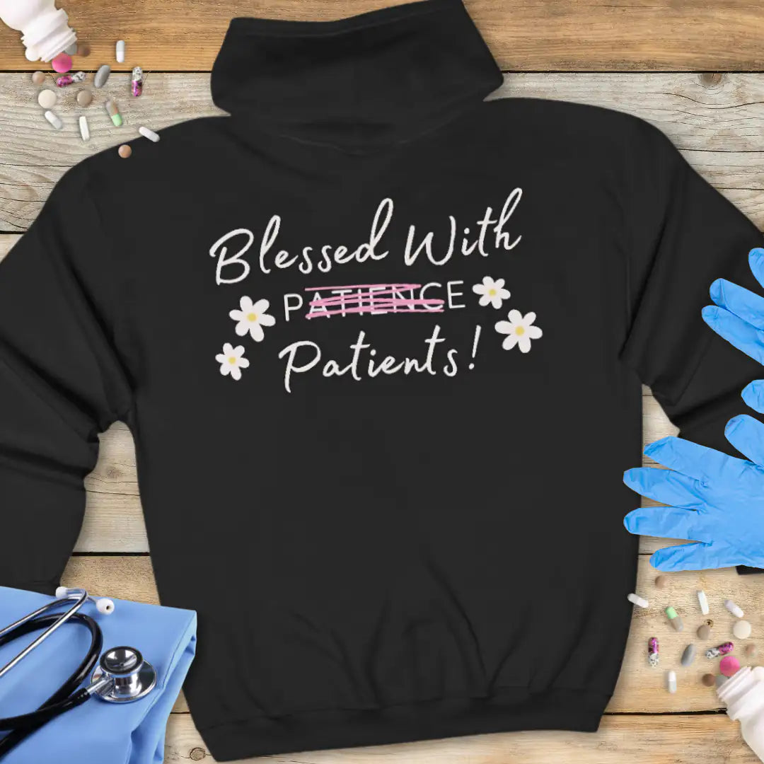 Black hoodie back with white script 'Blessed With Patients!' and a strikethrough 'Patience' in pink lettering, surrounded by white daisy graphics, on a wooden background with medical props.