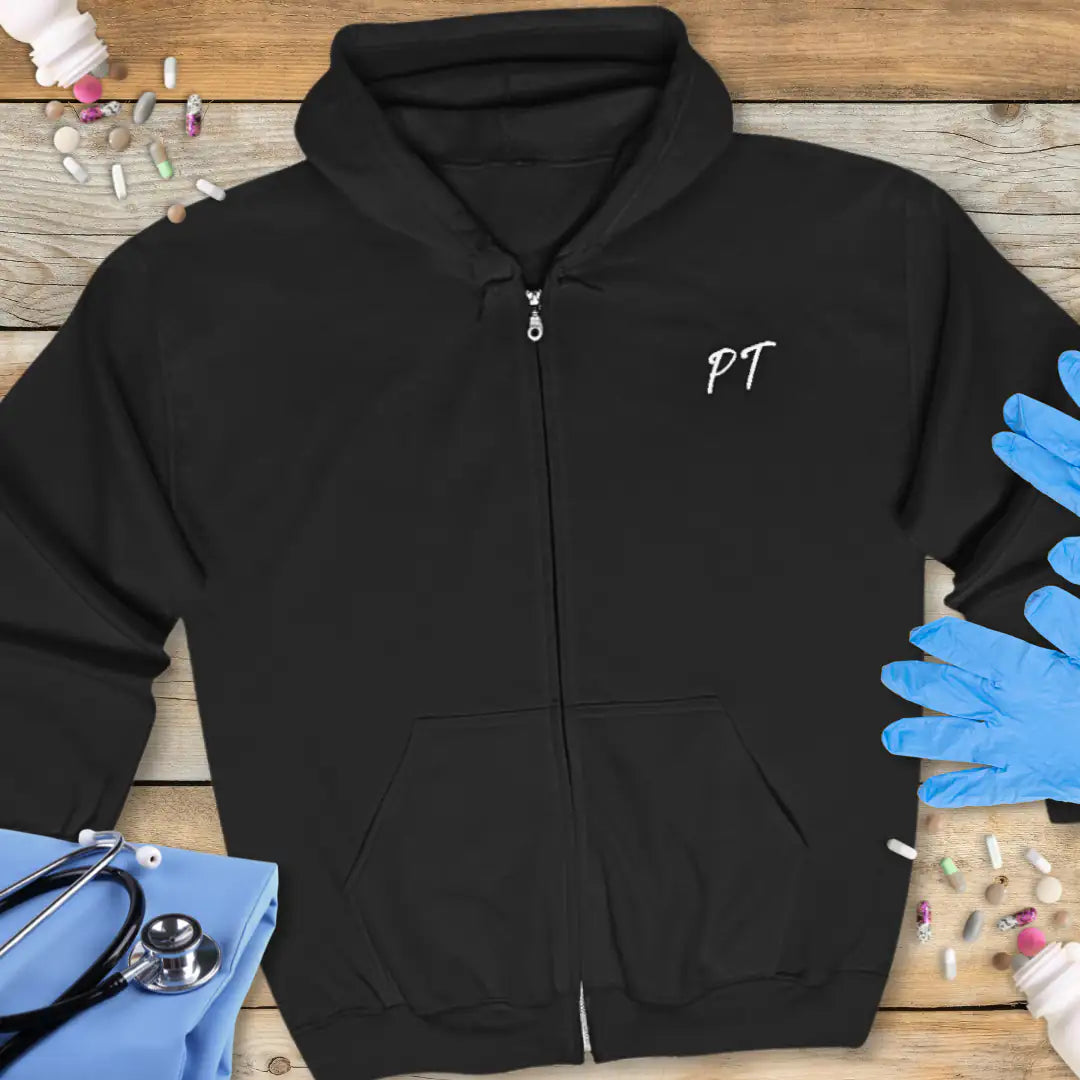 Black zip-up hoodie front with small white 'PT' script initials on the chest, displayed flat on a wooden background with stethoscope and medical gloves.