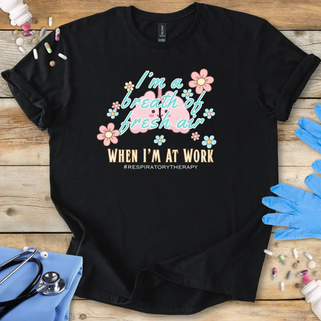  Black t-shirt with lung cartoon character and 'I'm a breath of fresh air when I'm at work' text on a wooden surface with medical supplies.