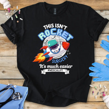 Black t-shirt with a humorous medical-themed graphic and 'This isn't rocket surgery' text on a wooden surface with medical accessories.