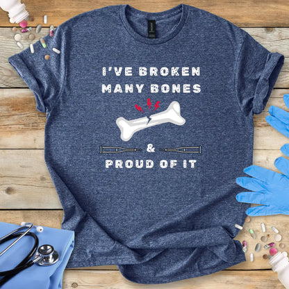 Blue med t-shirt with humorous text' I have broken many bones and proud of it' print on a wooden surface with medical accessories.