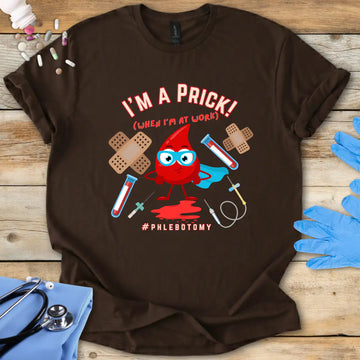 Brown t-shirt with a graphic design featuring a blood drop character and text 'I'm A Prick Phlebotomy' on a wooden surface with medical accessories.