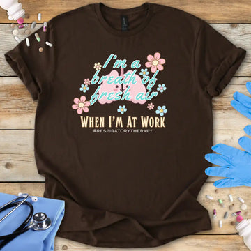 Brown t-shirt with lung cartoon character and 'I'm a breath of fresh air when I'm at work' text on a wooden surface with medical supplies.