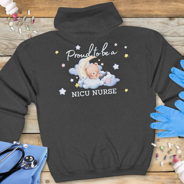 Dark grey hoodie back with white 'Proud to be a NICU Nurse' text and a cute teddy bear on clouds with moon and stars illustration, on a wooden background with stethoscope and medical gloves.