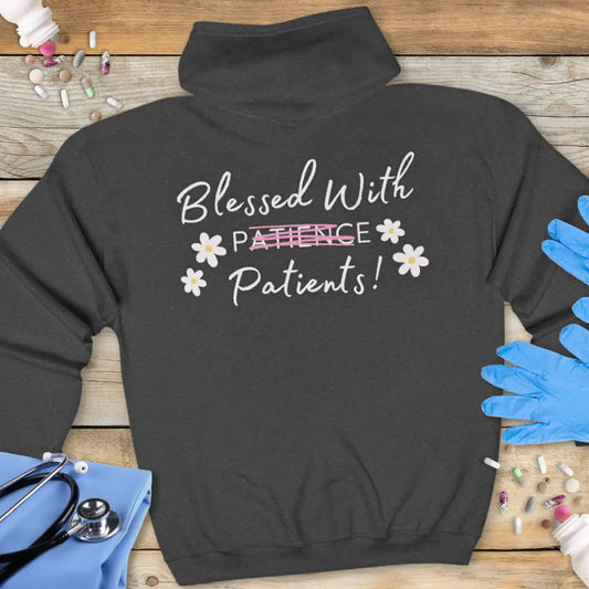 Dark grey hoodie back with 'Blessed With Patients!' in handwritten-style white text, with 'Patience' crossed out in pink and four white daisy accents, on a wooden background with stethoscope and gloves.