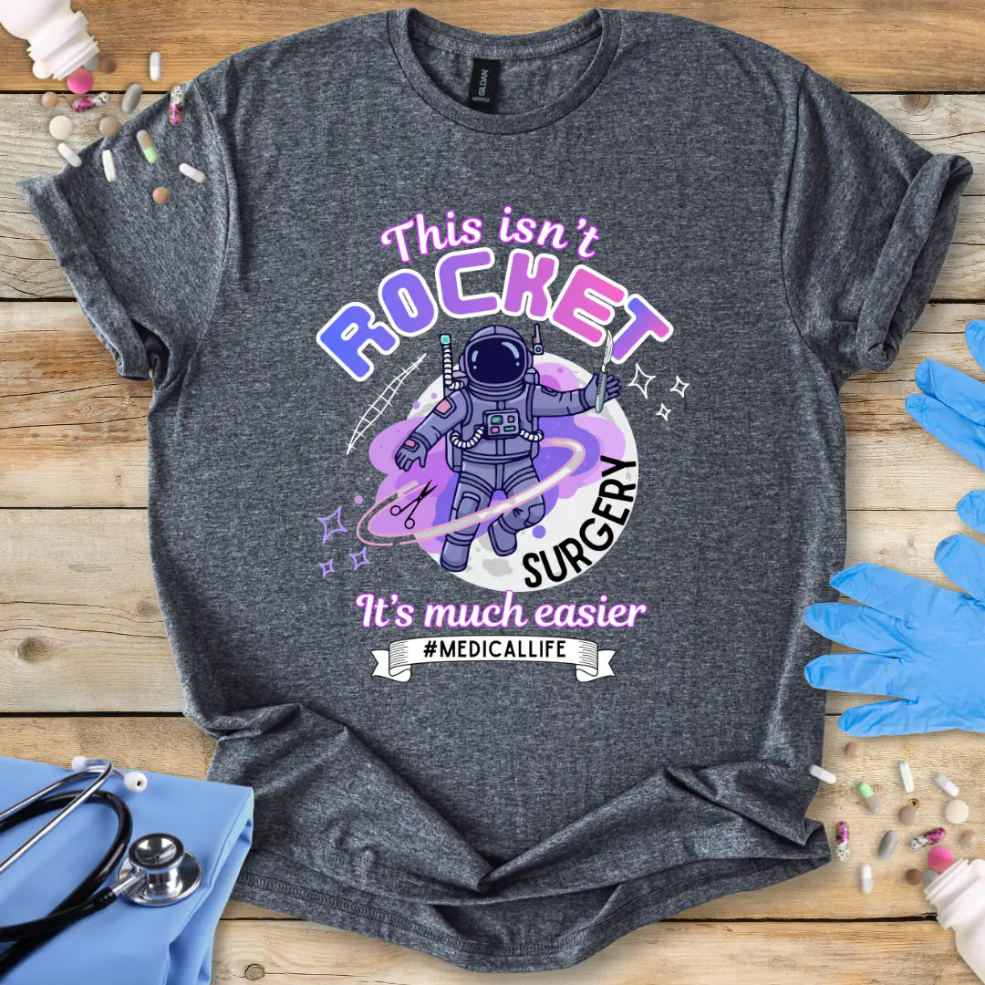 Gray t-shirt with space-themed graphic and text 'It's Not Rocket Surgery' on a wooden surface with medical supplies.