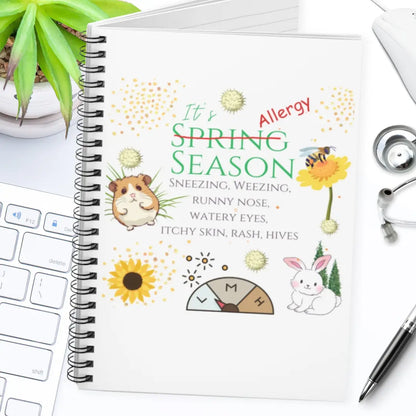 Notebook with allergy-themed cover and 'It's Allergy Season' text on a desk with a laptop and stethoscope.