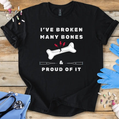 Black med t-shirt with humorous text' I have broken many bones and proud of it' print on a wooden surface with medical accessories.