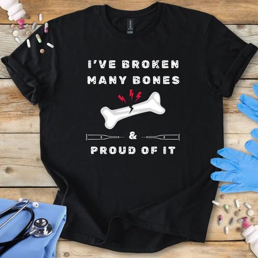 Black med t-shirt with humorous text' I have broken many bones and proud of it' print on a wooden surface with medical accessories.