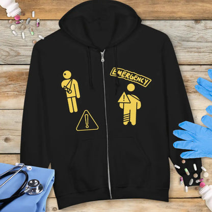 Black zip-up hoodie with yellow 'Emergency' graphic featuring injured patient stick figures and warning triangle icons on a wooden background with stethoscope and medical gloves.