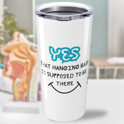 White tumbler with 'Yes That Hanging Ball Is Supposed To Be There' text and a blue graphic on a blurred background