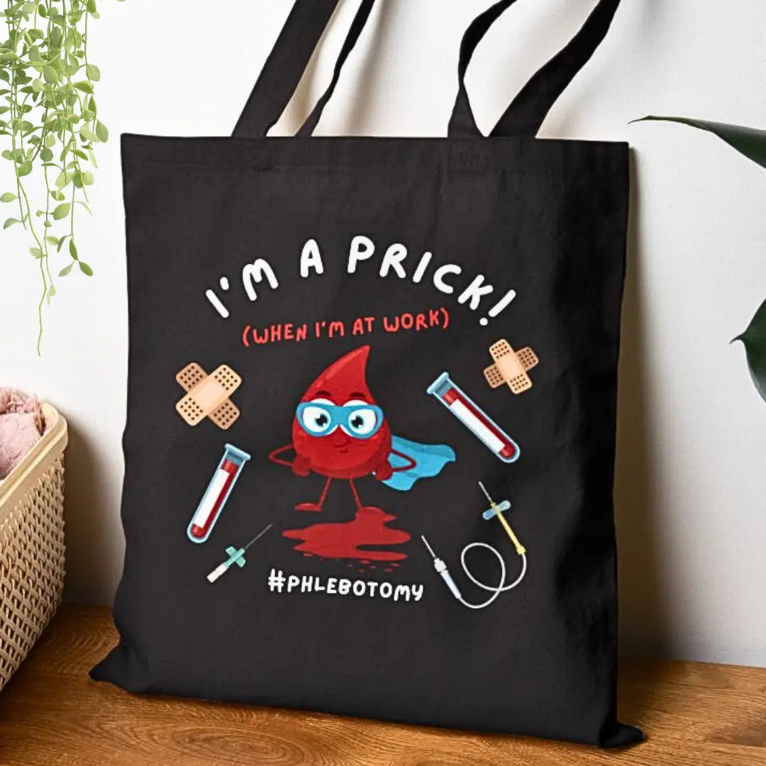 Black medical tote bag with a red cartoon blood drop and 'I'm A Prick When I'm At Work' text on a wooden surface.