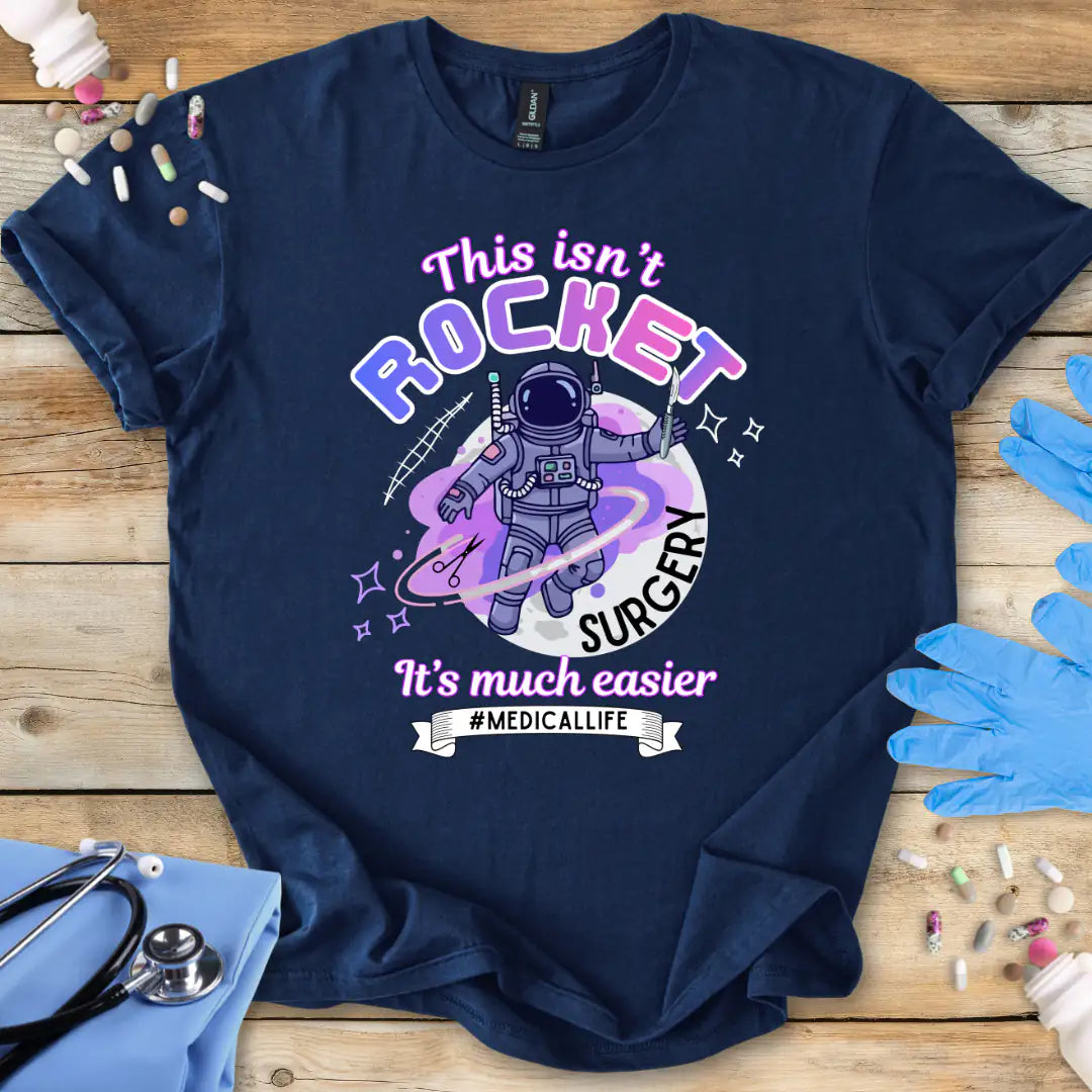 Navy blue t-shirt with space-themed graphic and 'This Isn't rocket surgery' text on a wooden surface with medical supplies.