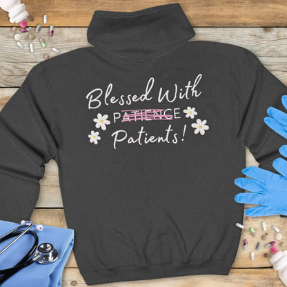 Dark grey hoodie back with 'Blessed With Patients!' in handwritten-style white text, with 'Patience' crossed out in pink daisy accents, on a wooden background with stethoscope.