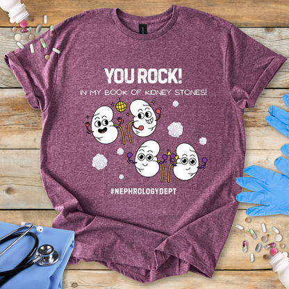 Heather Maroon gildan t-shirt with cartoon kidneys and 'You rock in my book of kidney stones, nephrology dept.' text on a wooden surface with medical accessories.