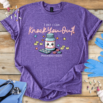 Heather purple t-shirt with a pharmacy-themed graphic and 'I bet I can knock you out pharmacy department' text on a wooden surface with medical accessories.