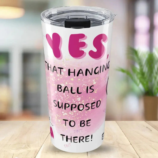 Pink tumbler with humorous text  'Yes, That Hanging Ball Is Supposed To Be There' on a blurred indoor background