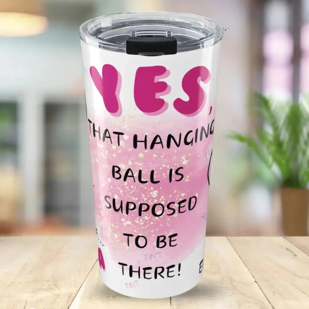 Pink tumbler with humorous text  'Yes, That Hanging Ball Is Supposed To Be There' on a blurred indoor background