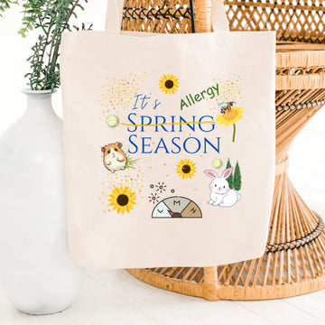 Cream canvas tote bag printed with 'It's Allergy Season' with 'Spring' crossed out in blue text, surrounded by sunflowers, pollen dots, a bee, a bunny, a hamster, and an allergy meter graphic, on a wicker chair background.