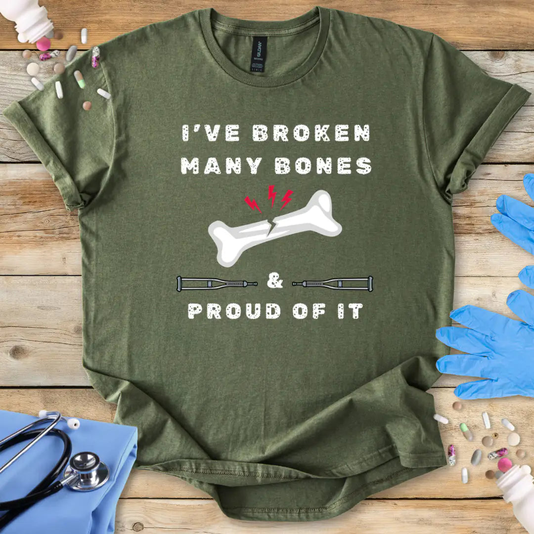 Military green med t-shirt with humorous text' I have broken many bones and proud of it' print on a wooden surface with medical accessories.