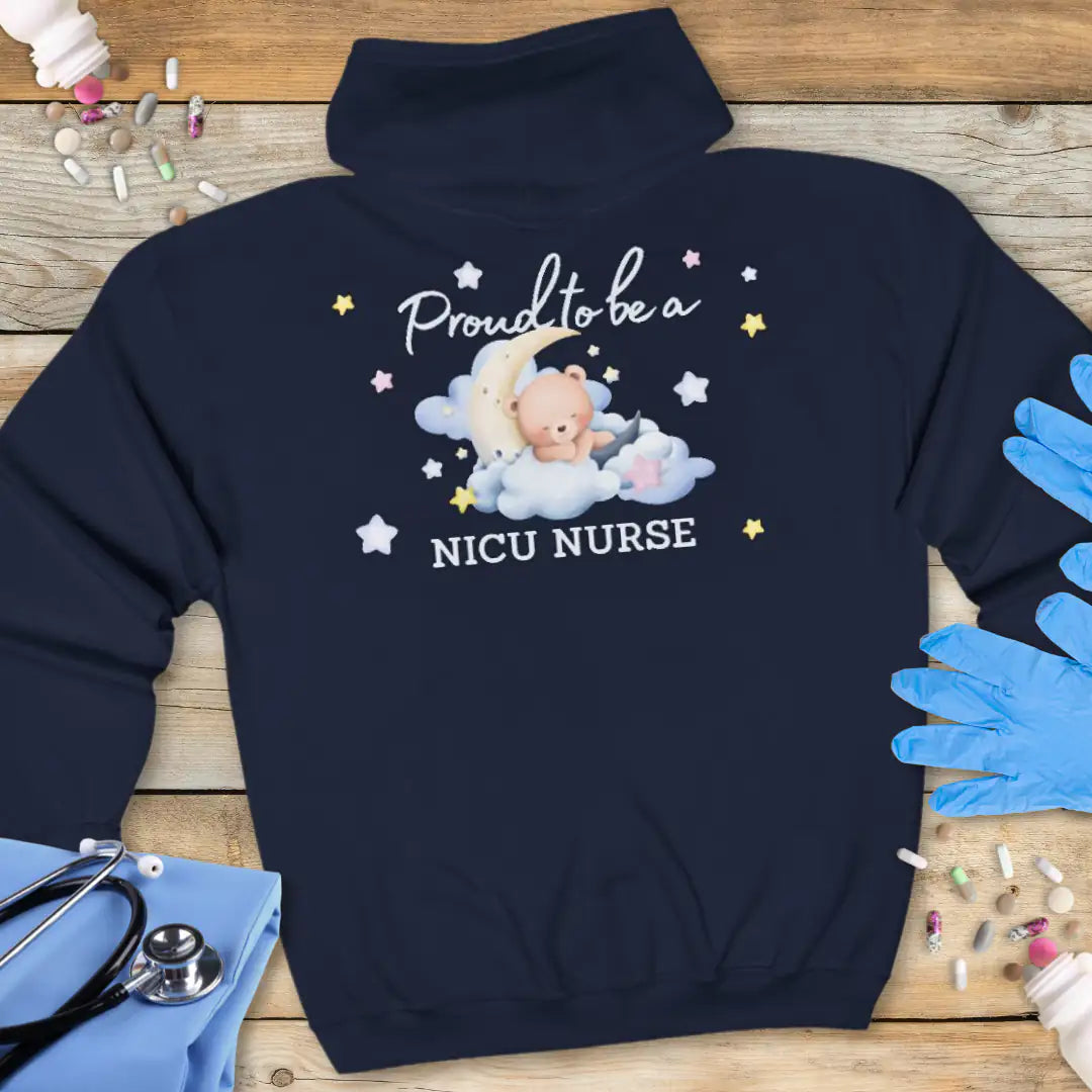 Navy blue hoodie back printed with 'Proud to be a NICU Nurse' in script and bold text, featuring a watercolor teddy bear resting on clouds with a crescent moon and colorful stars, on a wooden background with medical props.
