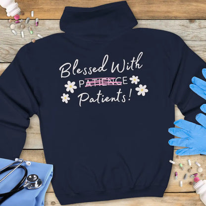 Navy hoodie back with 'Blessed With Patients!' in script text, featuring 'Patience' crossed out in pink with white daisy flowers scattered around, on a wooden background with stethoscope and medical gloves.