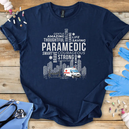 Navy blue t-shirt with paramedic-themed text and graphics on a wooden surface with gloves and an ambulance toy.