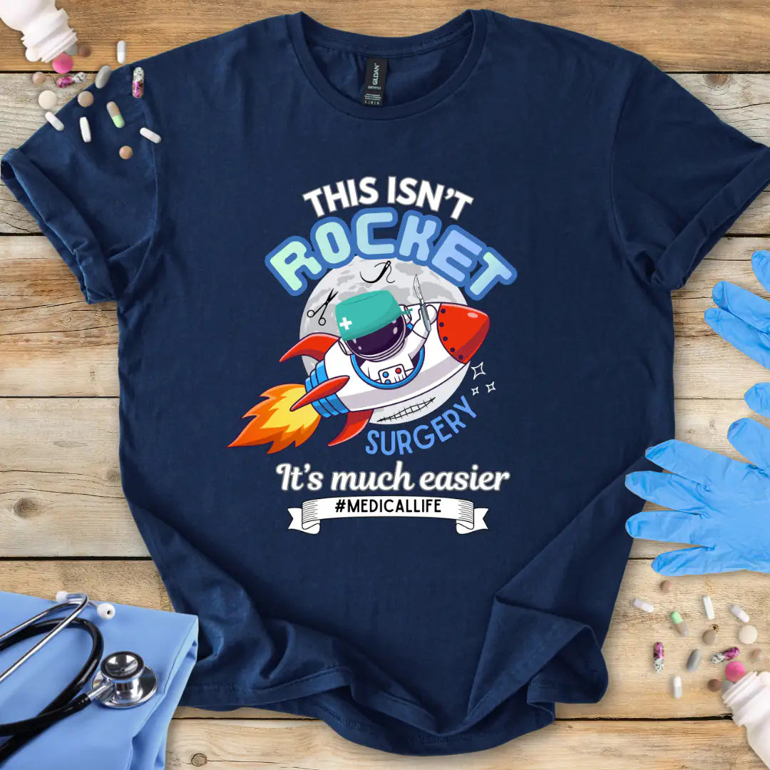 Navy blue t-shirt with a humorous medical-themed graphic and 'This isn't rocket surgery' text on a wooden surface with medical accessories.
