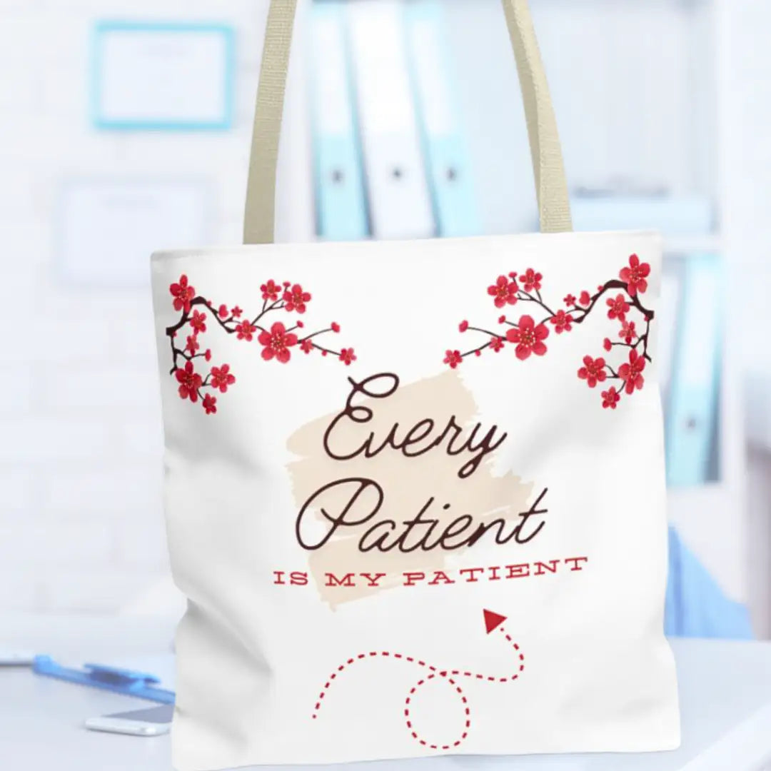 White canvas tote bag with beige straps printed with 'Every Patient Is My Patient' script text, red cherry blossom branches across the top, and a red dotted stethoscope illustration, on a blurred office background.