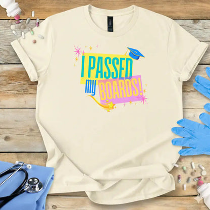 Beige t-shirt with 'I Passed My Boards!' text on a wooden surface with medical-themed items.
