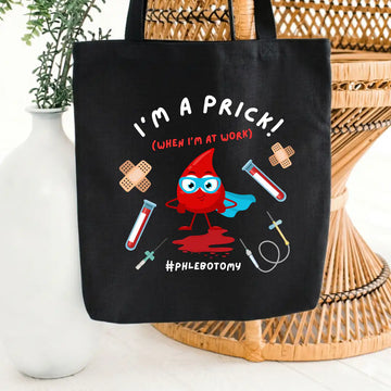 Black canvas tote bag printed with 'I'm A Prick! (When I'm At Work) #Phlebotomy' in white and red text, featuring a superhero blood drop character surrounded by blood vials, syringes, and bandage graphics, on a wicker chair background.