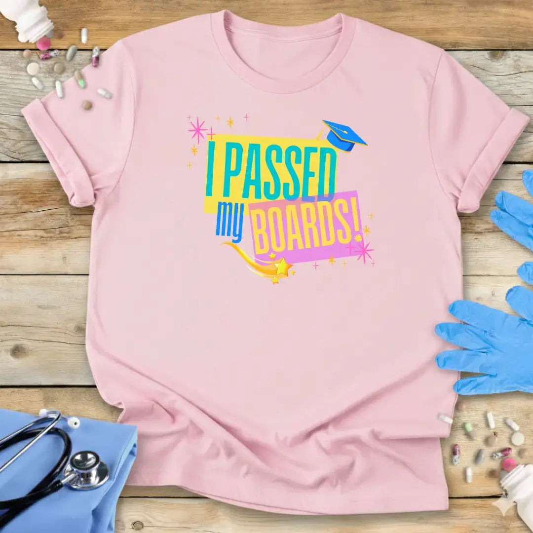 Pink t-shirt with 'I Passed My Boards!' text on a wooden surface with medical items.