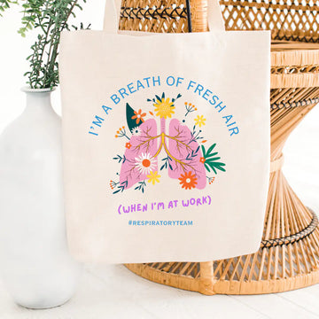 Cream canvas tote bag printed with 'I'm A Breath Of Fresh Air (When I'm At Work) #RespiratoryTeam' in blue and purple text, featuring a floral lung illustration with colorful wildflowers, on a wicker chair background.