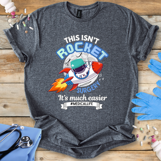 Gray t-shirt with a humorous medical-themed graphic and 'This isn't rocket surgery' text on a wooden surface with medical accessories.