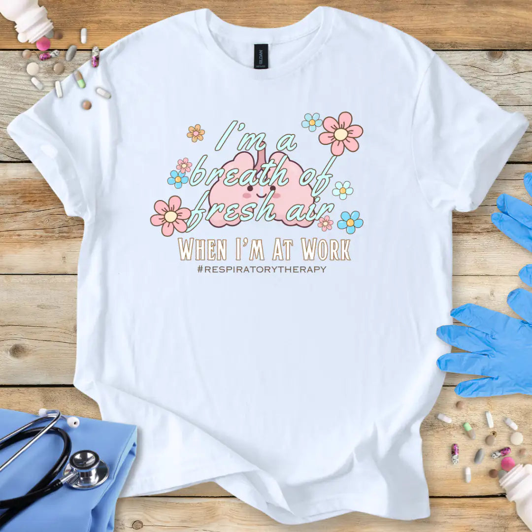 White t-shirt with lung cartoon character and 'I'm a breath of fresh air when I'm at work' text on a wooden surface with medical supplies.